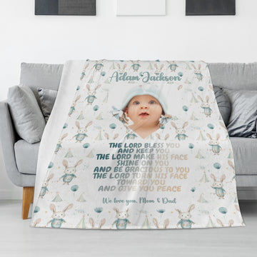 A Personalized Baby Name Blanket is elegantly placed over a gray couch, showcasing an image of a baby along with charming bunnies, balloons, and a blessing text. The blanket is customized with the baby's name at the top and includes a loving message from the parents at the bottom.