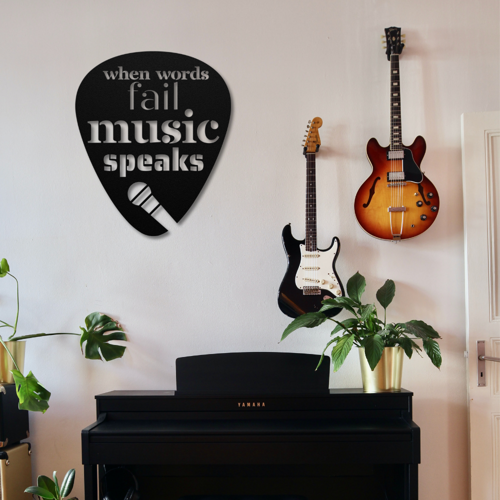 A music-themed room showcases a black digital piano beneath a guitar pick-shaped powder-coated metal sign inscribed with "When Words Fail Music Speaks | Mic." Above, two guitars hang on the wall, complemented by potted plants perched on the piano.