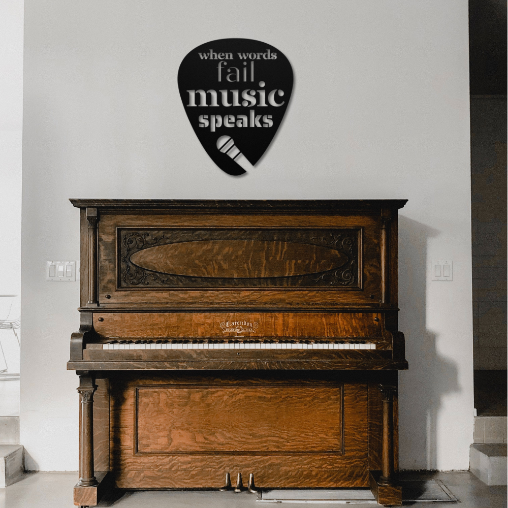 A vintage wooden piano stands against a white wall, beautifully accented by the "When Words Fail Music Speaks | Mic" sign. This powder-coated metal sign, shaped like a guitar pick and featuring the phrase along with a small microphone graphic, adds charm to your home decor.