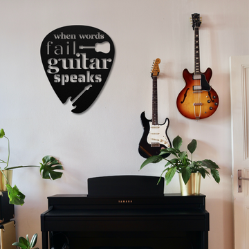 A room features a black upright Yamaha piano adorned with green plants in gold pots. A powder-coated metal sign from the "When Words Fail Guitar Speaks" collection, shaped like a guitar pick, adds an artistic touch. Enhancing the home's decor, two guitars—one sunburst and one black electric—are mounted on the wall.