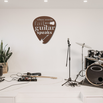 A minimalistic stage setup features a microphone, electric guitar, and drum set. A metal sign in the shape of a guitar pick displays the product name "When Words Fail Guitar Speaks." With plants in the corner, the ambiance is clean and modern, echoing stylish home decor.