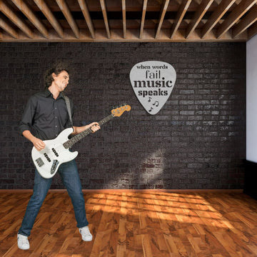 In a room with wooden flooring and a brick wall decorated with music-themed wall art, a man plays an electric guitar. The space features the "When Words Fail, Music Speaks" Guitar Pick Metal Wall Art, crafted from durable powder-coated steel in the USA, making it an ideal gift for guitarists. Wooden beams enhance the ceiling while sunlight creates patterns on the floor—truly an excellent setting for any Guitar Player gift.