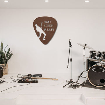 In a minimalist music studio, you'll find a drum set, microphone stand, and guitar on the floor. Adding charm above is the "Eat, Sleep, Play, Repeat" Guitar Pick Metal Wall Art made from durable powder-coated steel. This striking piece is perfect for guitarists and can be used indoors or outdoors. The studio also features a touch of green from a potted plant in the corner.