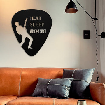 Above a brown leather sofa adorned with gray pillows hangs the "Eat, Sleep, Rock, Repeat Guitar Pick Metal Wall Art." This piece, made from durable powder-coated steel in the USA, features a guitarist's silhouette and is an ideal gift for guitarists. A modern black lamp casts its glow over this versatile art piece suitable for both indoor and outdoor use.