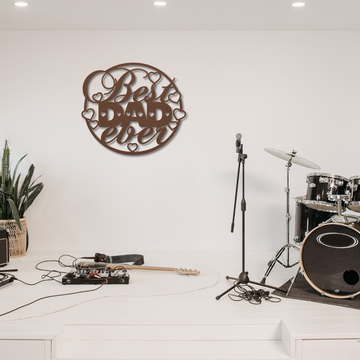 A minimal stage setup with a drum set, two microphones, guitars on the floor with cables and pedals, a green potted plant, and a laser cut metal wall art sign that reads "Best Dad Circle Sign.