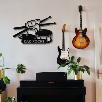 A black upright piano faces a white wall with two electric guitars and a “Dad Rocks” metal wall art featuring a hand, drumsticks, hihat, and snare drum above it; green potted plants finish the music room decor.