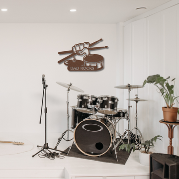A black drum set and microphone stand sit in a bright music room. Above, the "Dad Rocks Sign with Hand, Drumsticks, Hihat and Snare Drum | Metal Wall Art" hangs proudly. A nearby plant adds a fresh touch to the modern decor.