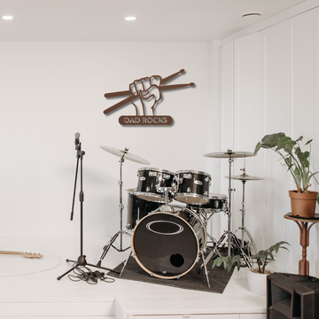 A black drum set sits on a rug next to a mic stand and guitar in a white room. A plant rests on a stool, and the Dad Rocks Sign with Hand and Drumsticks | Metal Wall Art adds perfect dad wall art or a great gift for dad.