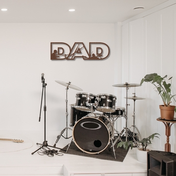 A black drum set and microphone stand sit on stage near a potted plant. Above, the "Dad Text Sign with Drummer Figures | Metal Wall Art" hangs on the white wall. A guitar rests on the stage floor.
