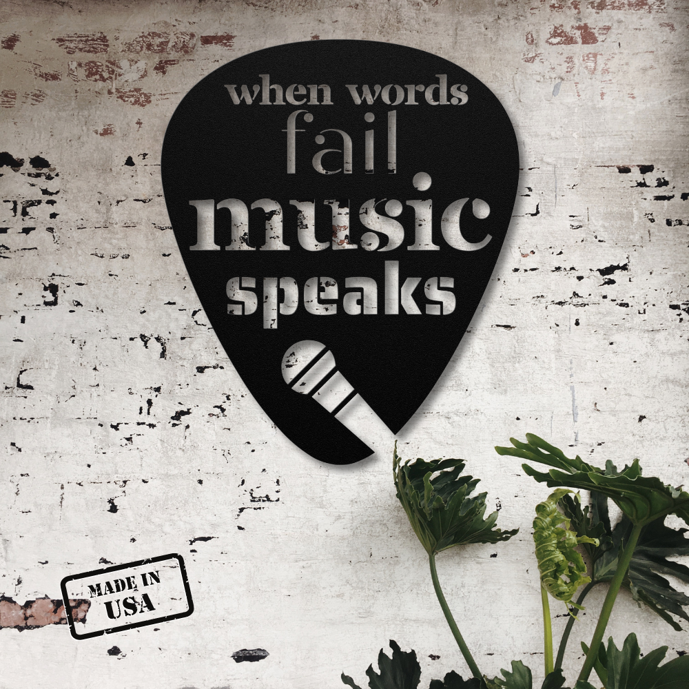 A black, guitar pick-shaped metal sign featuring the phrase "when words fail, music speaks" is mounted on a white brick wall. A small plant adds a touch of greenery in the lower right corner. This piece of home decor, named "When Words Fail Music Speaks | Mic," is powder-coated and stamped with "Made in USA" in black on the bottom left side.