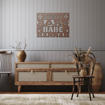 A rustic living room showcases a wooden sideboard, ceramic vase with dried branches, wooden chair, and the "Badass Bass Babe | CUSTOM Metal Wall Art for Female Bass Guitarist" hung in cutout letters above the sideboard.