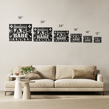 A modern living room features a beige sofa and six metal wall art pieces in various sizes (12"–36"), each reading "badass BABE NANA" with florals—perfect as Badass Bass Babe custom wall art for female bass guitarists.