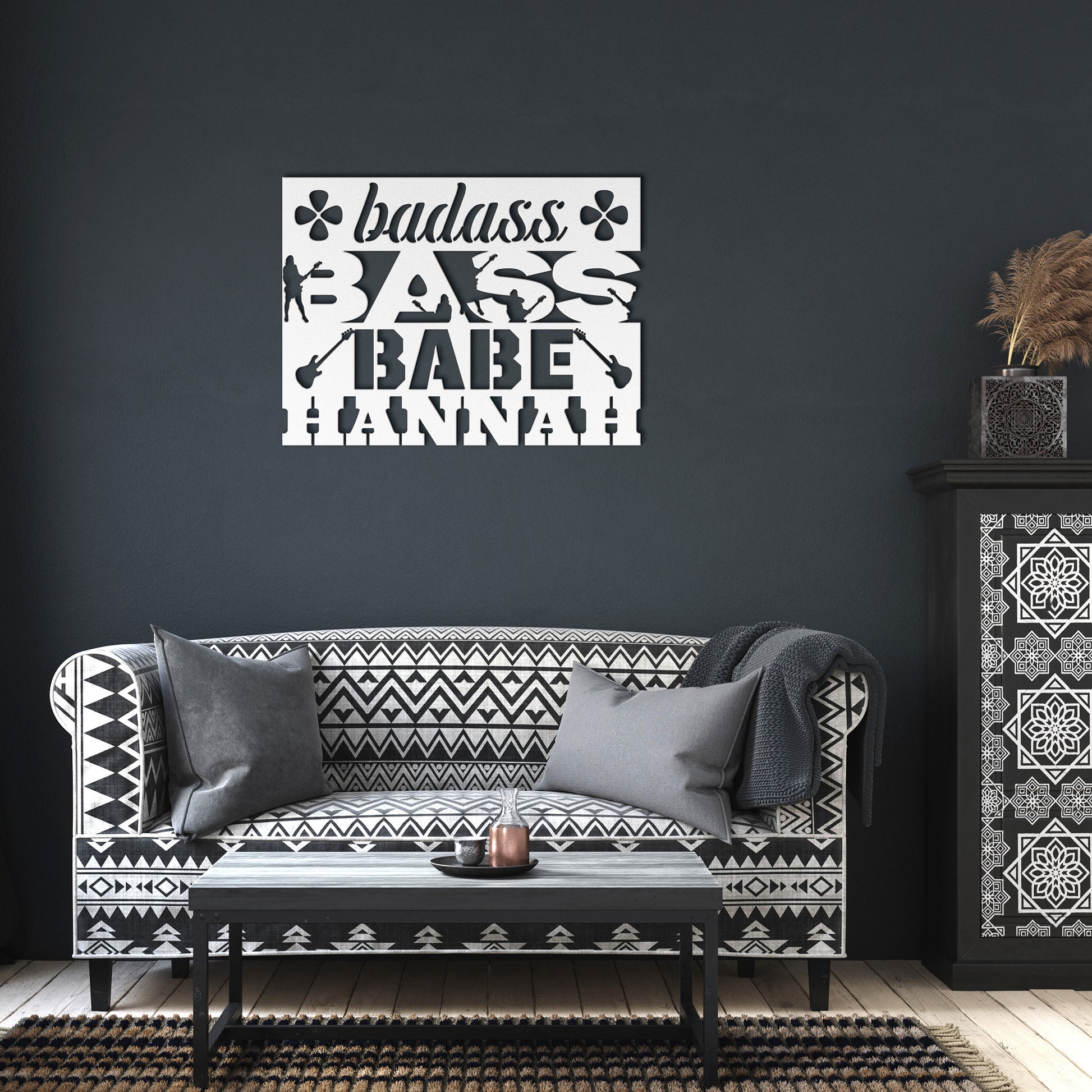 A black and white living room features a patterned sofa, matching table, and dresser with vases. Above the “badass BASS BABE HANNAH” wall sign hangs the Badass Bass Babe | CUSTOM Metal Wall Art for Female Bass Guitarist.