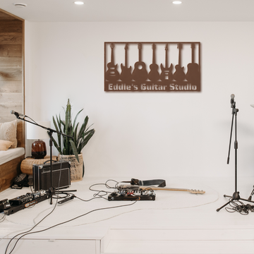A music studio with guitars, amps, and mics features the Guitar Wall Sign Custom Name—custom metal wall art reading “Eddie’s Guitar Studio” with guitar silhouettes above. Plants and cushions decorate a corner of the white floor room.
