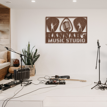 A stylish home music studio setup features a Guitarist Singer Wall Sign Custom Name | Custom Metal Wall Art above plants, microphones, a guitar, speakers, and cables on a white floor with a cozy seating area for modern decor.