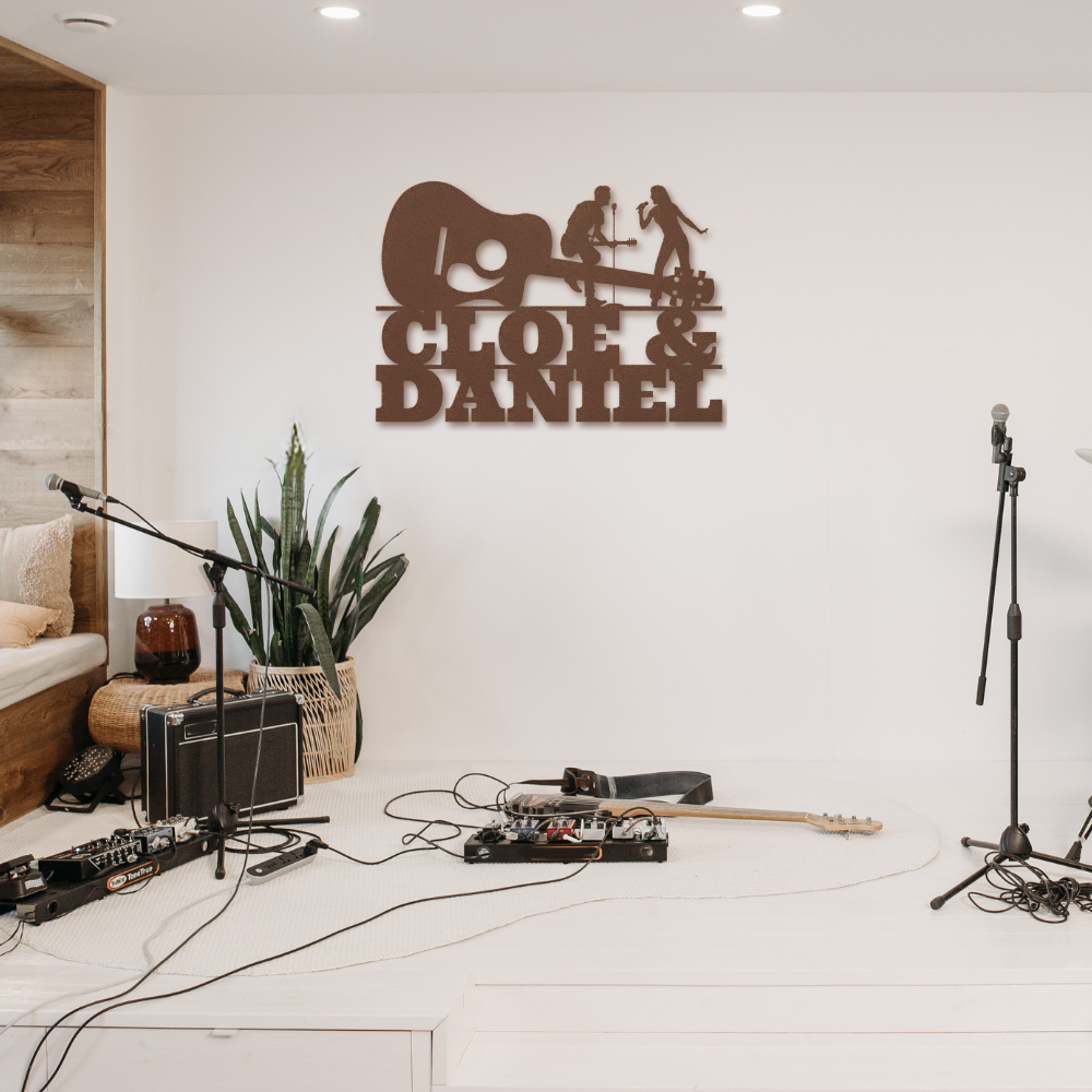 A stage features musical gear, mics, pedals, and amps. On the wall is the Singer & Guitarist Wall Sign with Custom Name (MWA-MCS-0122), a powder-coated metal art piece featuring a guitar silhouette and "Cloé & Daniel" in bold script. A potted plant adds decor.