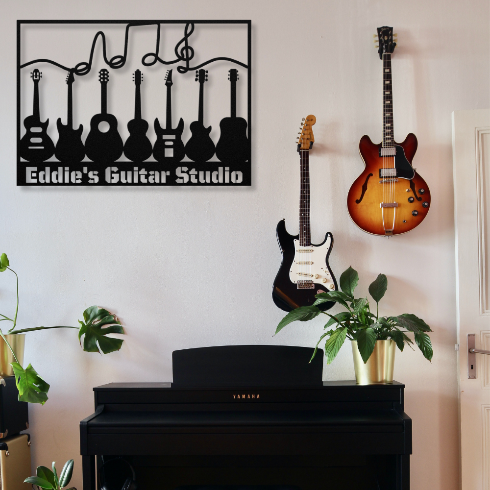 A custom metal wall sign, "Eddie's Guitar Studio," from the Multiple Guitars Wall Sign with Music Notes and Custom Text, hangs above a piano. Below it, black and sunburst guitars are mounted on a white wall with green plants on the piano.