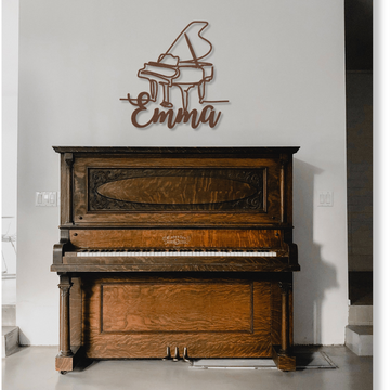 A vintage upright piano stands against a white wall. Above it, the "Grand Piano Line-Art with Custom Name" metal sign displays a grand piano outline and "Emma," creating unique, personalized decor in the well-lit, minimalist space.