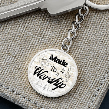 A "Made to Worship Gold Sheet Music" keychain for drummers, engraved with musical notes and "Made to Worship," is attached to a key and displayed on textured fabric—a perfect gift for any drummer.