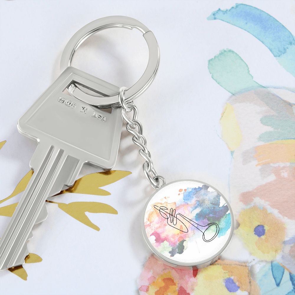 The Trumpet Colorful Circle Pendant Keychain features a vibrant watercolor background with a simple trumpet line drawing, attached to a silver key and keyring, displayed on abstract watercolor artwork. Ideal gift for trumpetists.