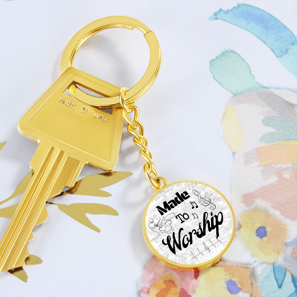 A silver key with a round, engraved "Made to Worship" keychain featuring drums, music notes, and sheet music—perfect for drummers—rests on an abstract floral-patterned background.