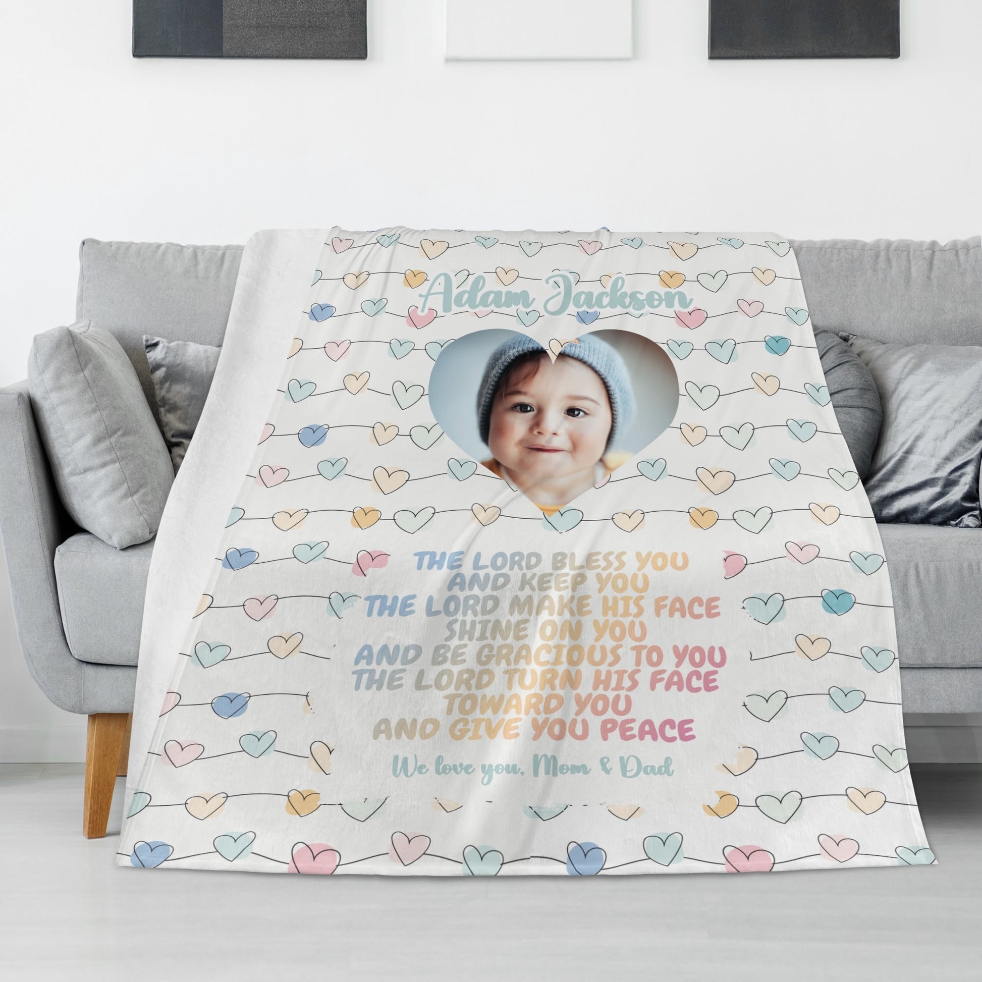 A cozy personalized baby name blanket featuring a heart-shaped cutout with a baby's face, adorned with colorful hearts and inscribed with the Bible verse: "The Lord bless you and keep you...". Signed, "We love you, Mom & Dad," this charming creation drapes beautifully over the couch. Perfect as a custom photo blanket gift for baptisms, baby showers, or Christmas.