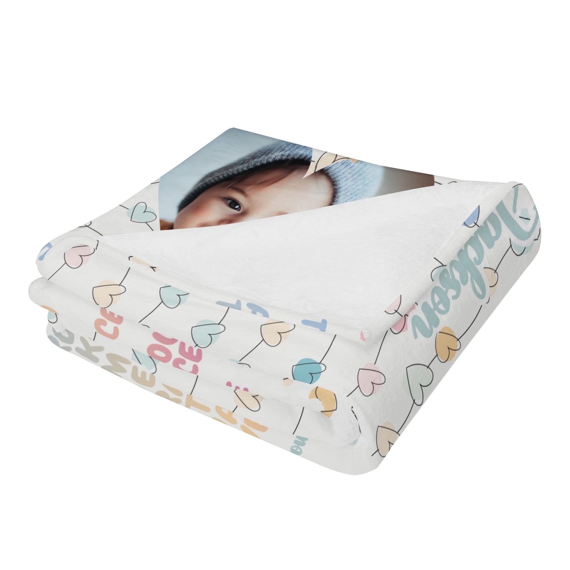 A folded white blanket adorned with colorful heart patterns and letters, showcasing a printed photo of a baby in a blue hat. The Personalized Baby Name Blanket beautifully blends warmth and love, making it an ideal baby blessing gift.