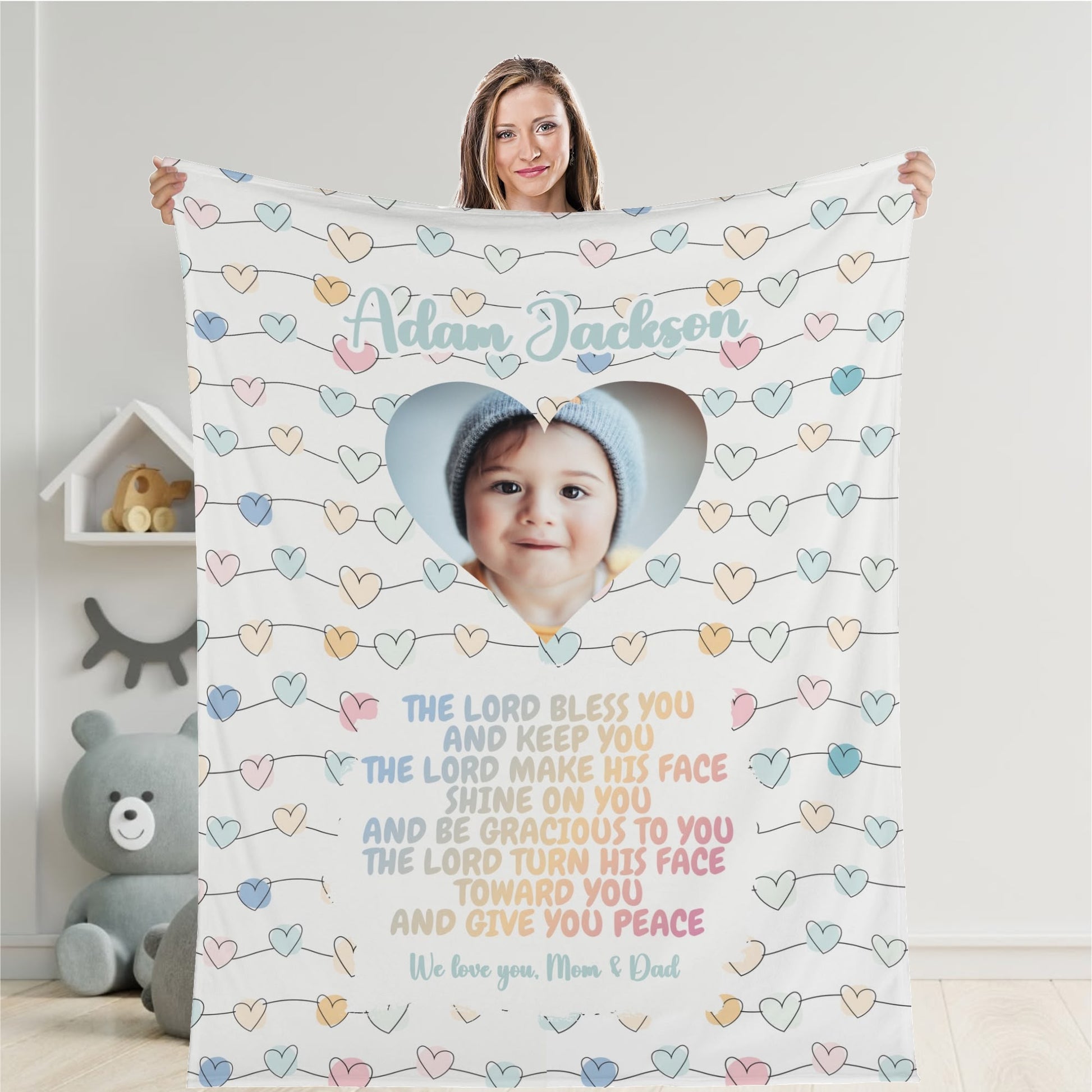 A person holds a Personalized Baby Name Blanket, featuring a baby's photo set in a heart-shaped frame. The image is encircled by pastel-colored hearts, with a blessing message below. "Adam Jackson" is printed at the top, making it a cherished keepsake for any nursery.