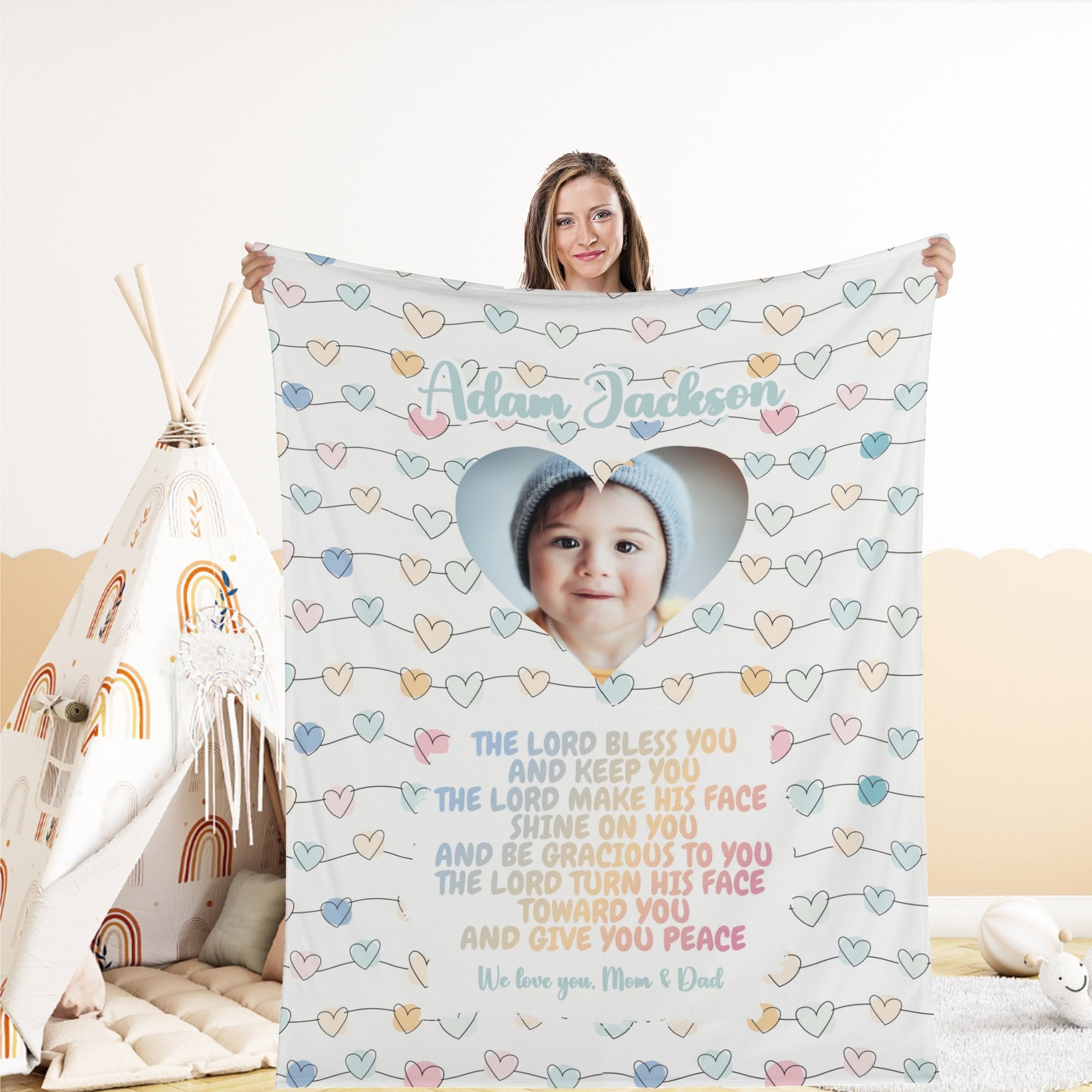 A woman holds a Personalized Baby Name Blanket, custom-designed with a child's photo framed by a heart shape. Decorated with pastel-colored hearts and featuring a prayer for blessing and peace, this customized newborn name blanket rests near a teepee adorned with rainbow patterns in the background.