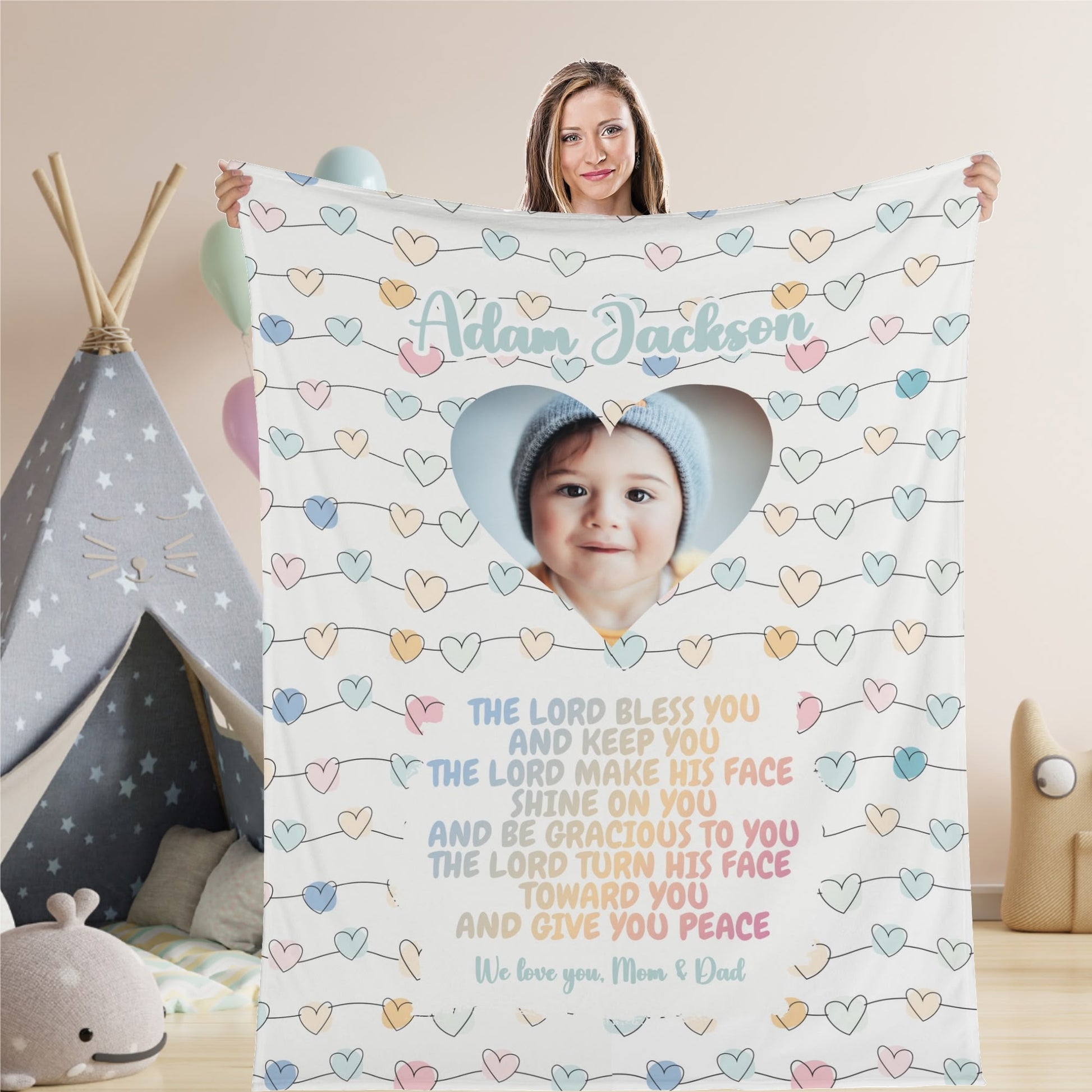 A woman holds a personalized baby name blanket showcasing the baby's smiling face within a heart-shaped cutout. The photo is surrounded by pastel-colored hearts and a blessing, accompanied by a personal message from "Mom & Dad." A canopy and toys in the background enhance the charm of this custom photo blanket gift, perfect for various occasions like baptisms, baby showers, or Christmas.