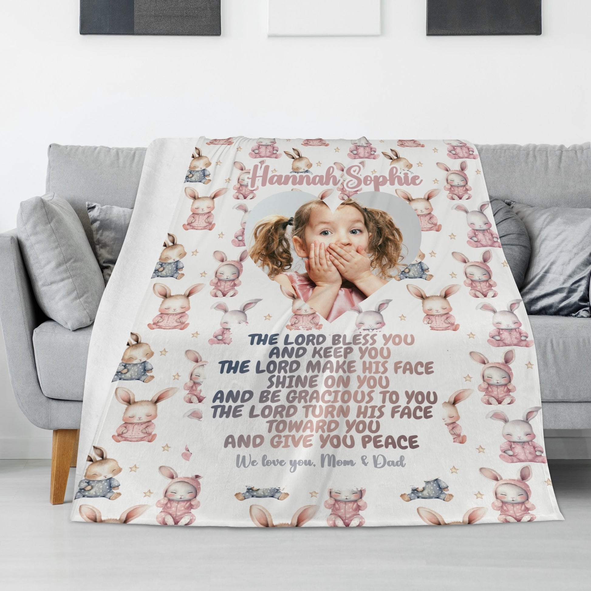 A Personalized Baby Name Blanket, adorned with pastel rabbits, is elegantly draped over a gray sofa. It showcases a child's photo alongside the heartfelt message: "The Lord bless you and keep you... The Lord turn His face toward you and give you peace. We love you, Mom & Dad.