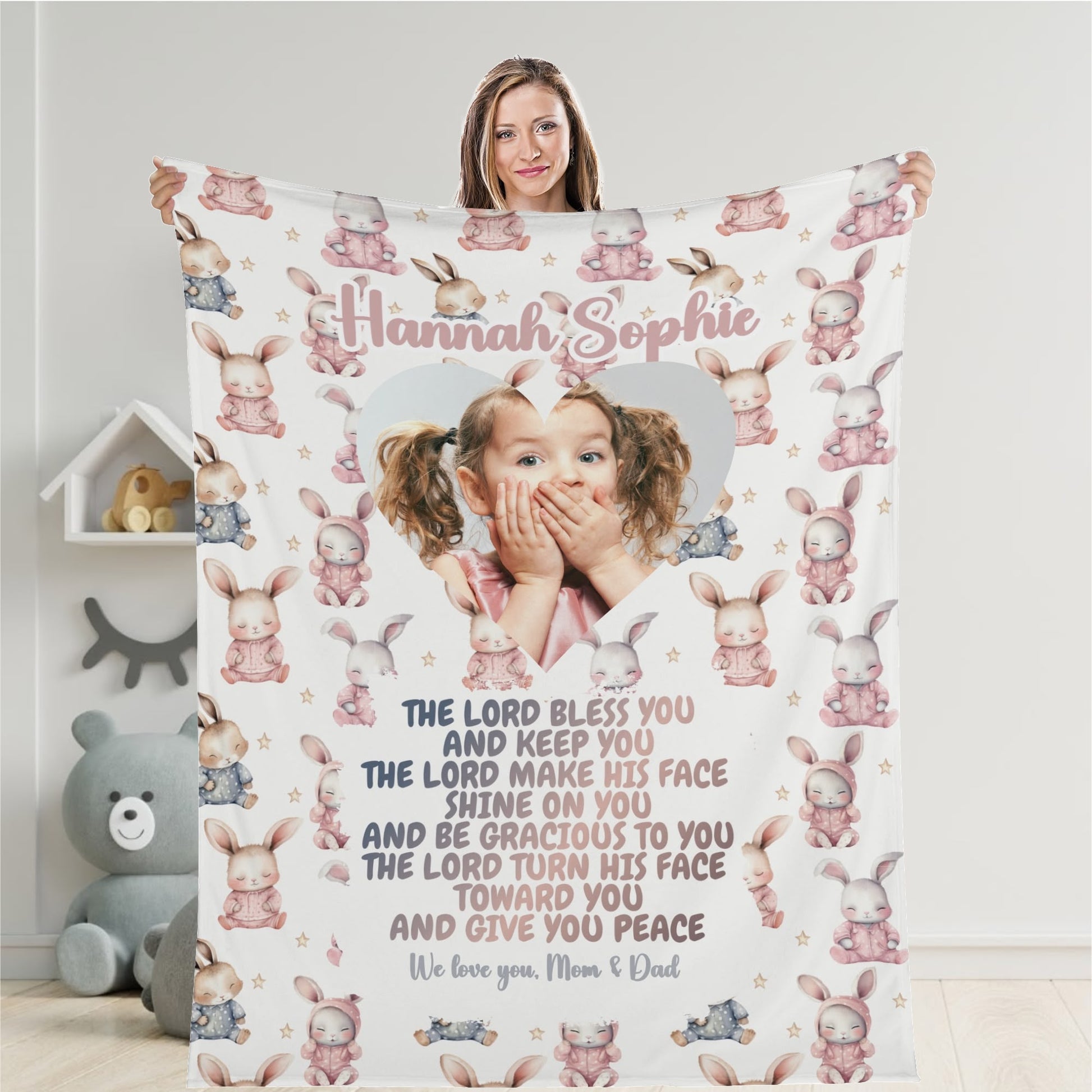 A woman holds a personalized baby name blanket customized with pastel-colored bunny designs and a heart-shaped cutout featuring an image of a child with hands on cheeks. The text includes a heartfelt message from "Mom & Dad" to "Hannah Sophie.