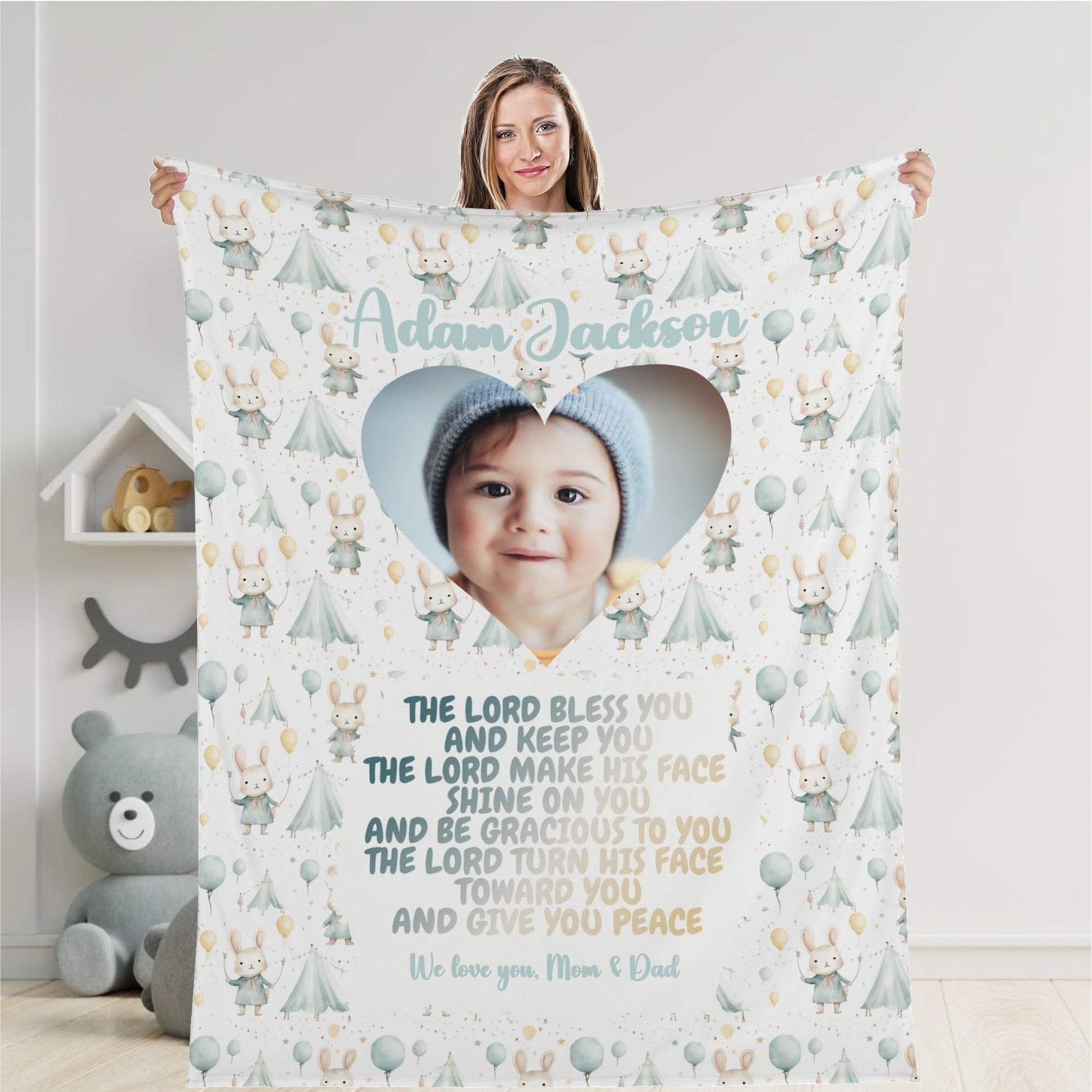 A woman holds a Personalized Baby Name Blanket that has been customized with a heart-shaped photo of a baby wearing a hat. The blanket features illustrations of tents and teddy bears, along with a heartfelt blessing and message from "Mom & Dad".