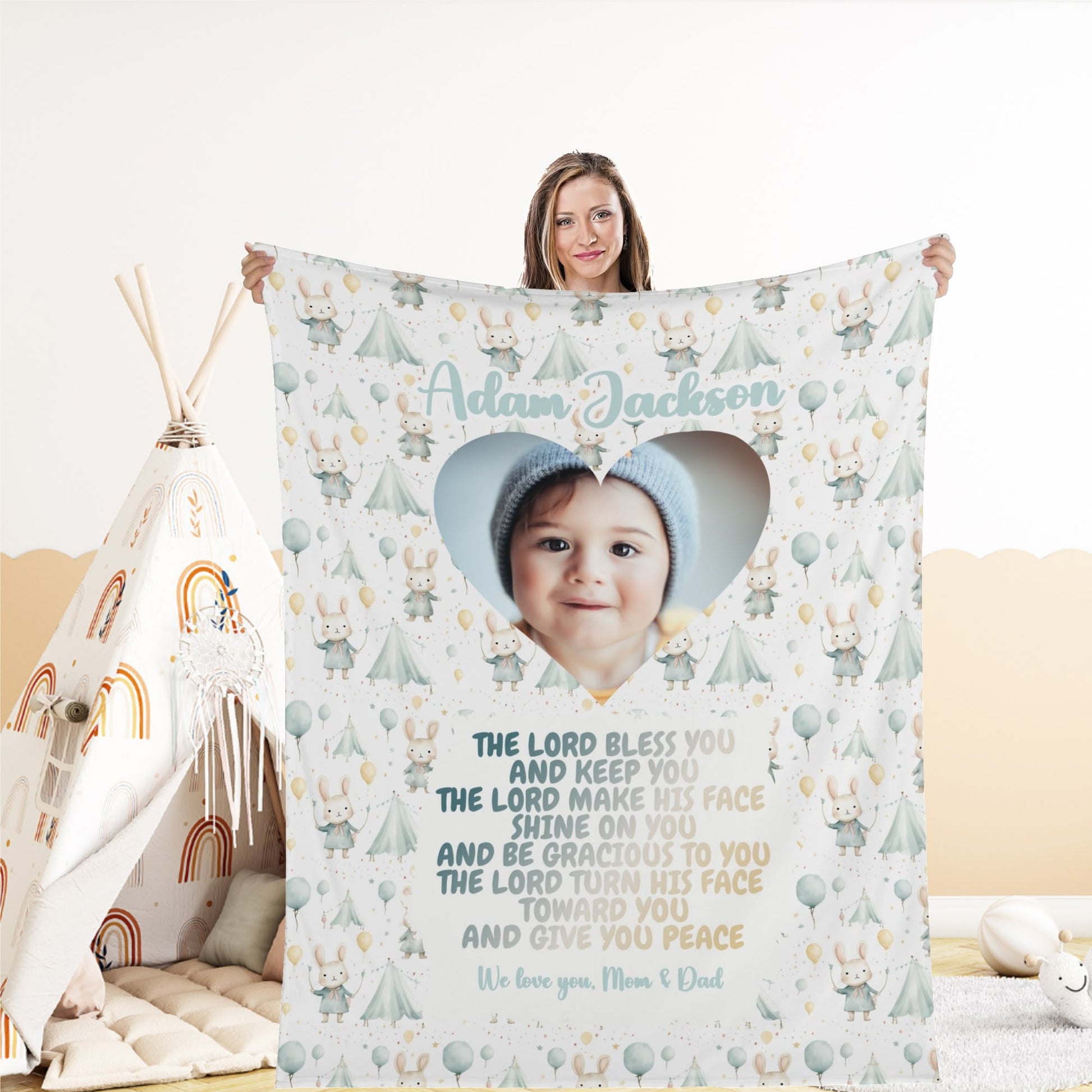 A woman holds a personalized baby name blanket featuring a heart-shaped image of a baby wearing a blue hat. This custom photo blanket gift is decorated with trees, bears, and balloons, accompanied by a heartfelt message from "Mom & Dad" and their chosen blessing.