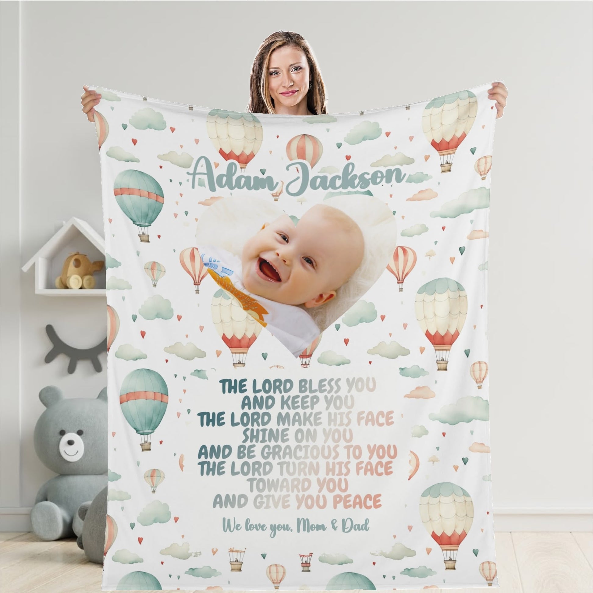 A smiling baby is wrapped in a Personalized Baby Name Blanket, customized with the name "Aidan Jackson." Featuring hot air balloons, clouds, and a prayerful Bible verse, this charming blanket is held by a woman standing beside a shelf adorned with a teddy bear and books.