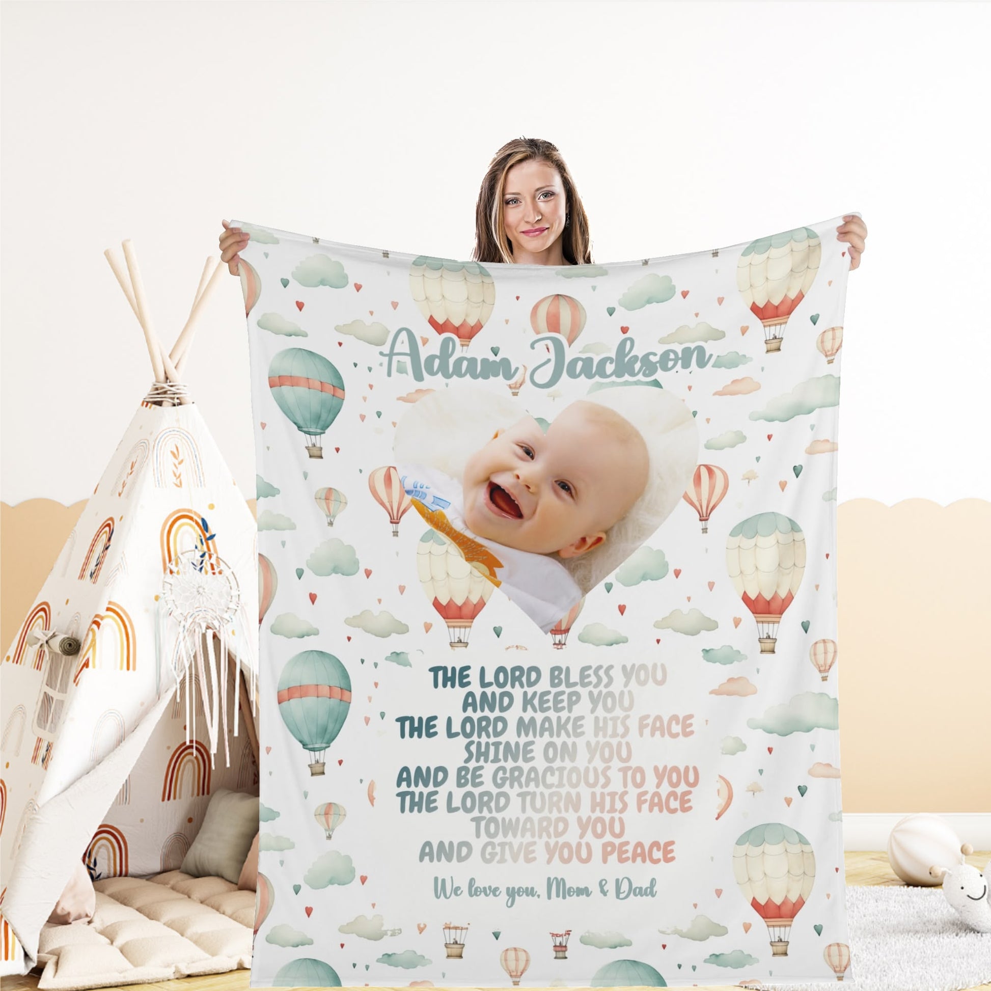 A woman holds a "Personalized Baby Name Blanket" featuring a cheerful baby, hot air balloons, and clouds. The customized text includes a Bible verse that starts with "The Lord bless you and keep you." A small teepee is seen in the background.