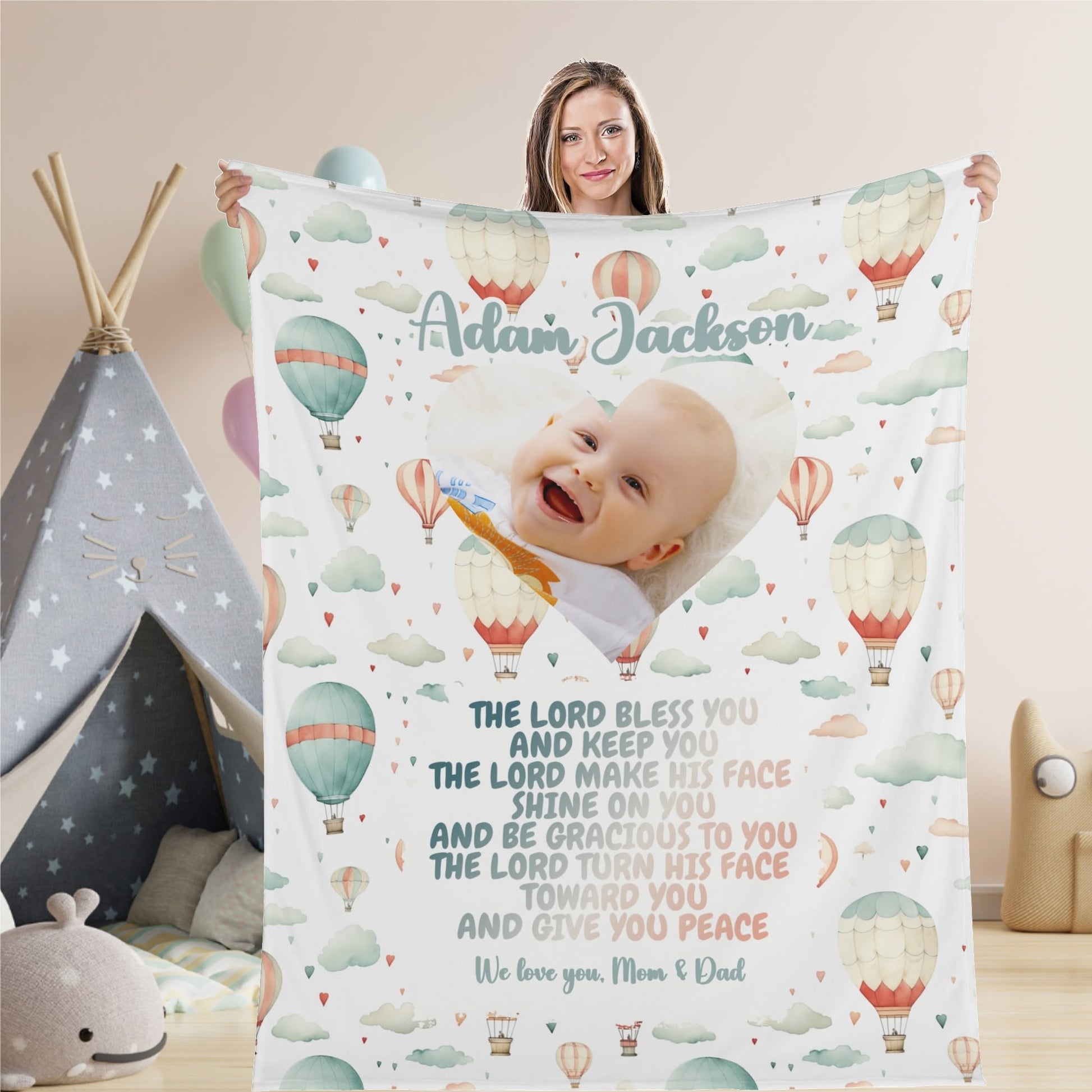 A smiling woman holds a Personalized Baby Name Blanket, featuring a happy baby, hot air balloons, clouds, and a heartfelt quote. The design conveys blessings, peace, and love from the parents, with "Adam Jackson" beautifully personalized at the top.