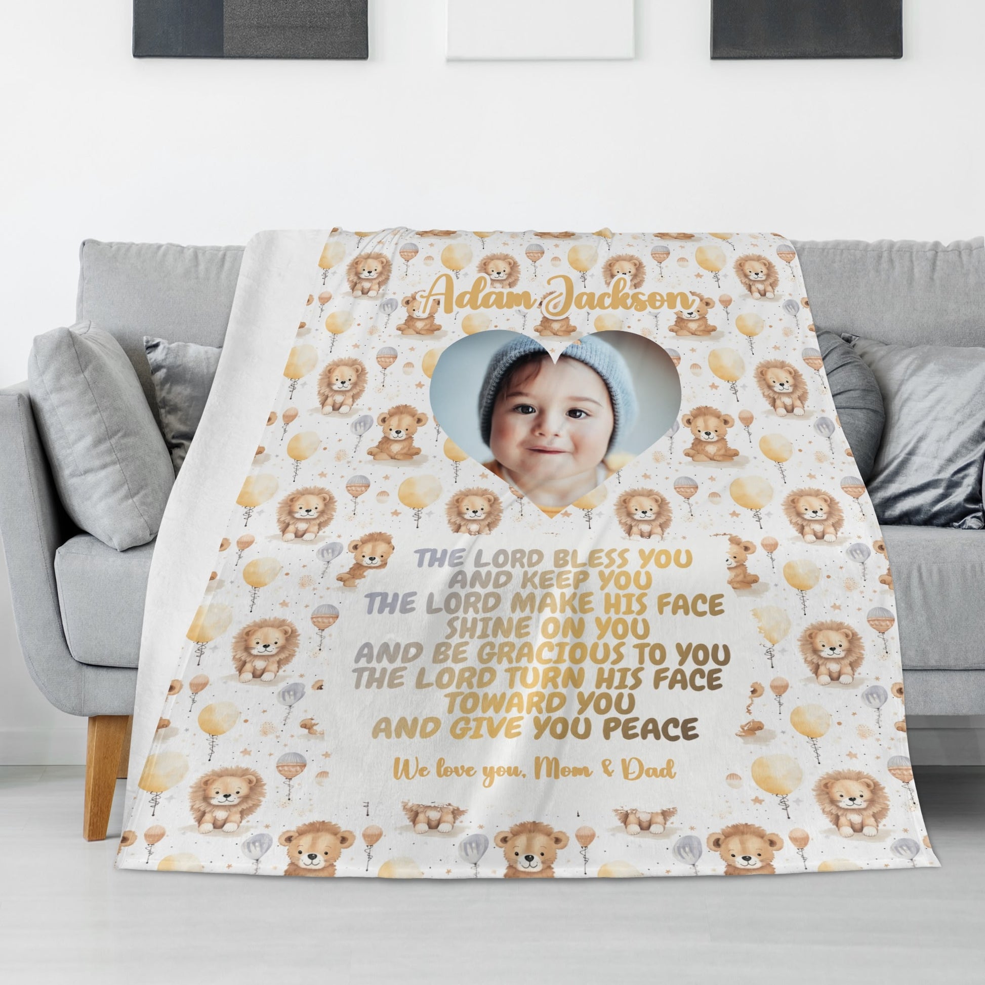 A Personalized Baby Name Blanket is beautifully draped over a sofa, showcasing adorable lion and balloon illustrations with a heart-shaped cutout displaying the baby's face. This Custom Photo Blanket Gift features personalized text with a heartfelt message from "Mom & Dad," accompanied by a tender blessing.