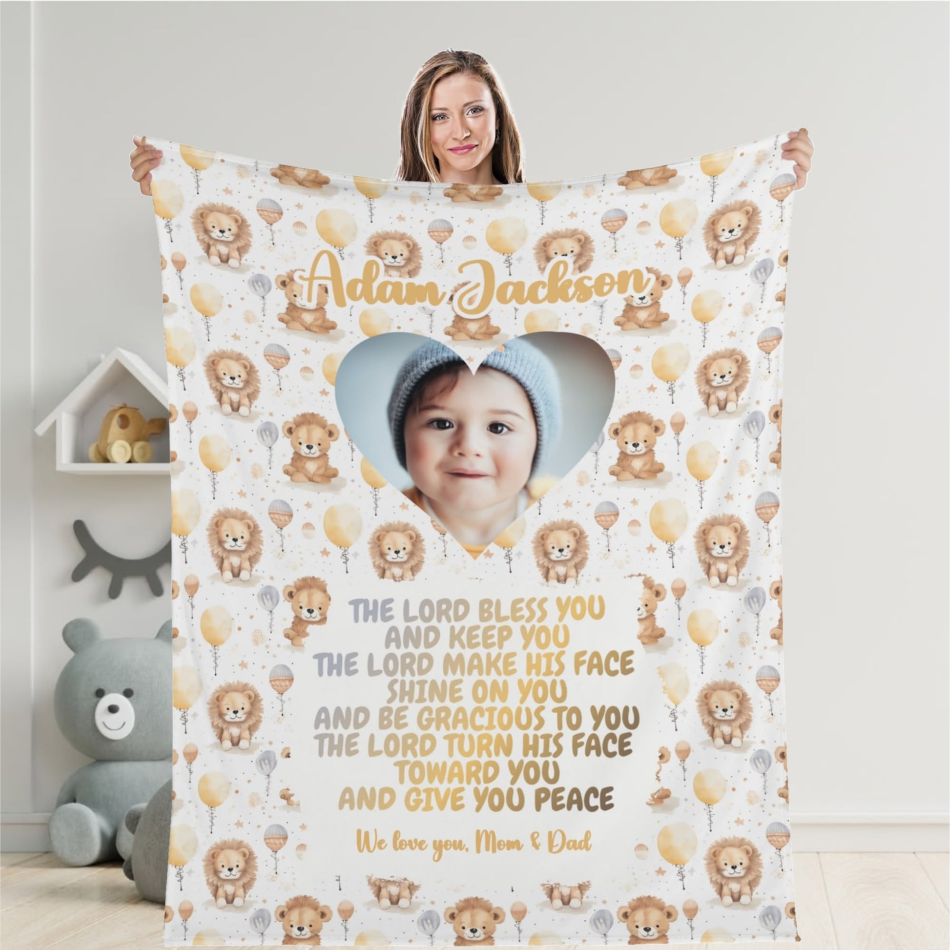 A woman holds a Personalized Baby Name Blanket, customized with a photo of a baby in a heart-shaped frame. Decorated with illustrations of lions and balloons, it features personalized text with a heartfelt blessing and the message "We love you, Mom & Dad.