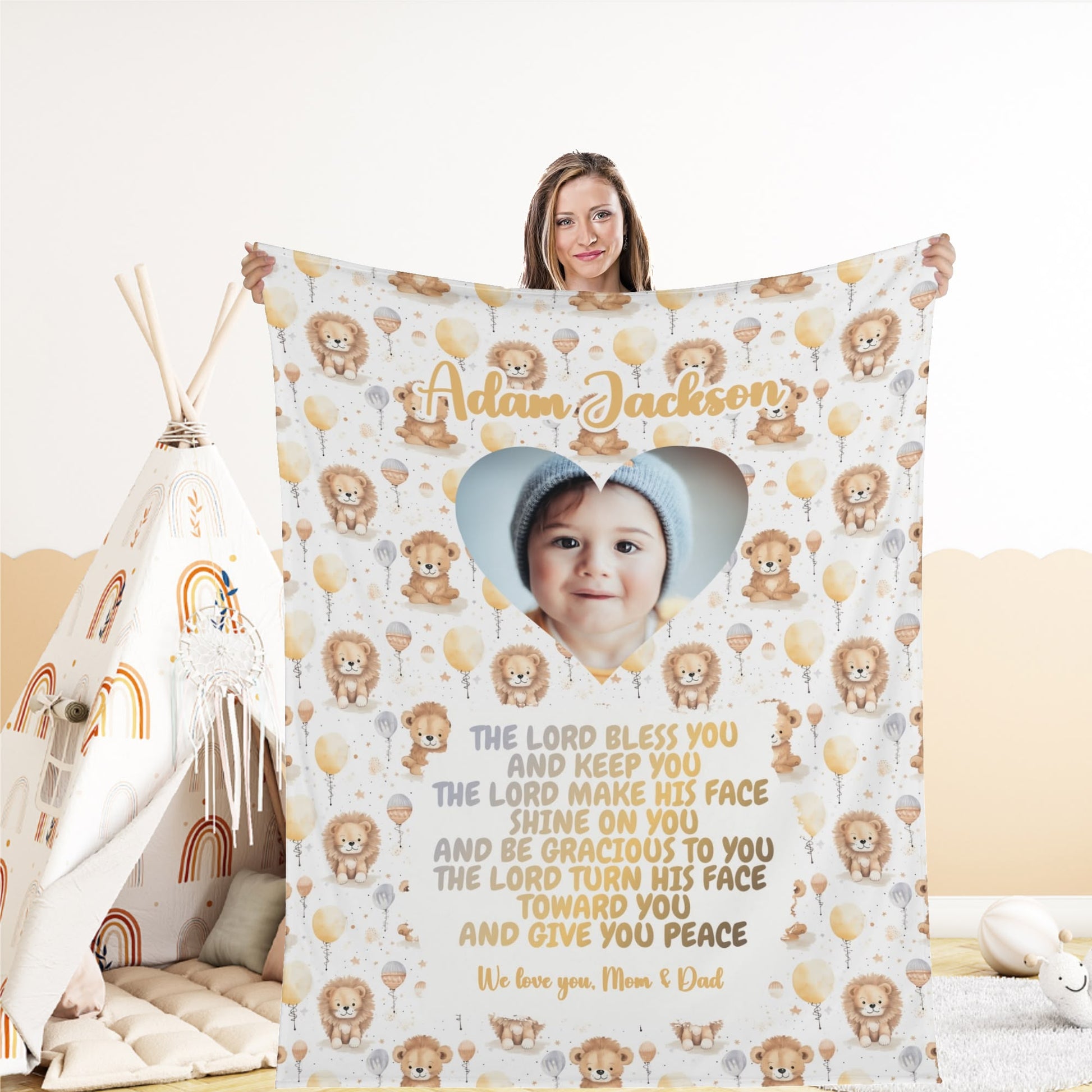 A woman holds a Personalized Baby Name Blanket adorned with a photo of a baby in a knitted hat. The blanket features lion and balloon illustrations alongside the text, "The Lord bless you and keep you..." followed by a loving message from "Mom & Dad.