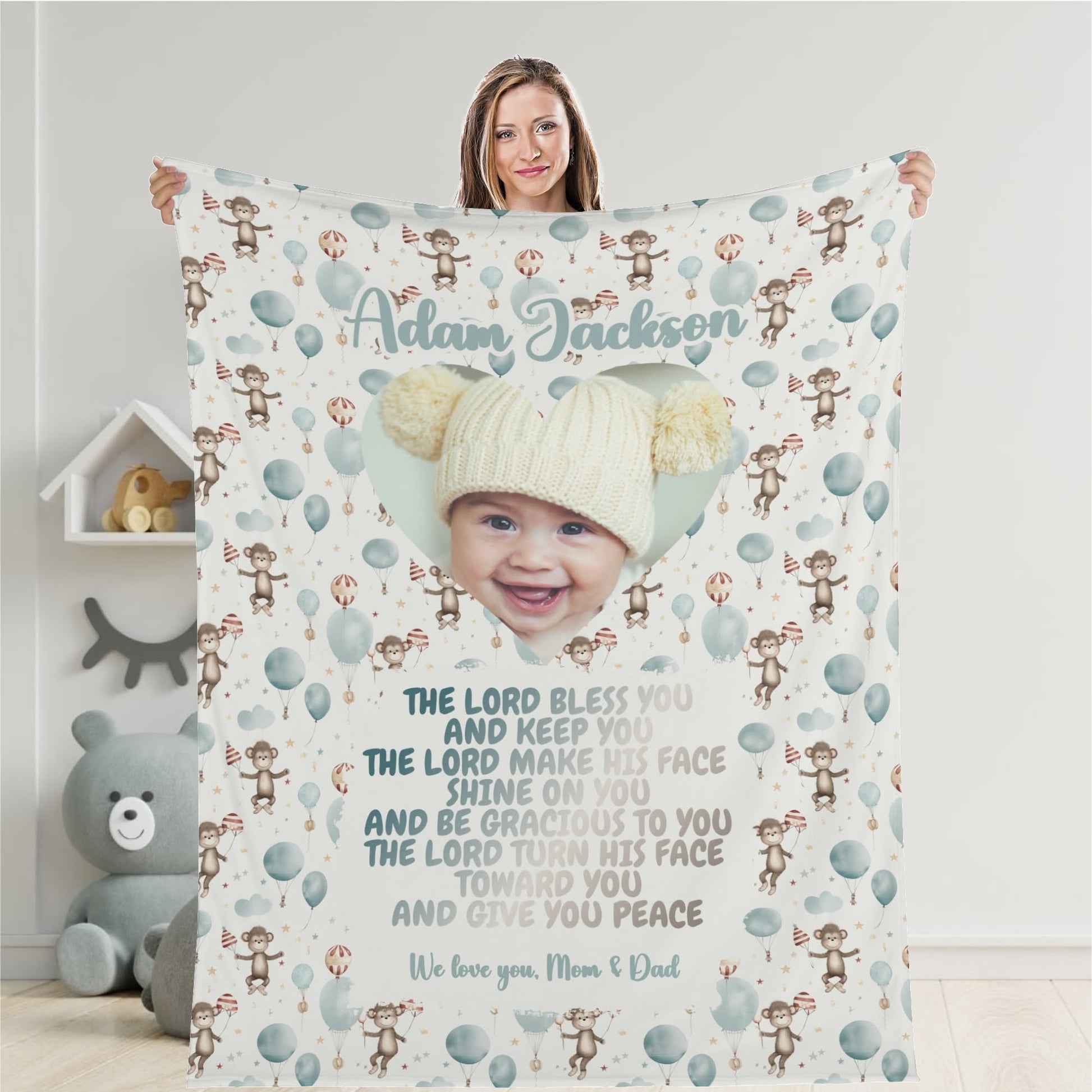 A woman holds a Personalized Baby Name Blanket, which has been customized to feature a baby's face in a heart shape surrounded by monkeys and balloons. The blanket includes a prayer along with the personalized text, "We love you, Mom & Dad.