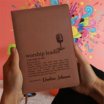 A person holds the “Personalized Worship Leader Journal – Vegan Leather Music Notebook” in brown, titled “worship leader,” with definitions, a microphone graphic, and personalized with the name Darlene Johnson. A pink wall adorned with colorful abstract art appears in the background—making this custom worship journal a thoughtful gift for Christian musicians.