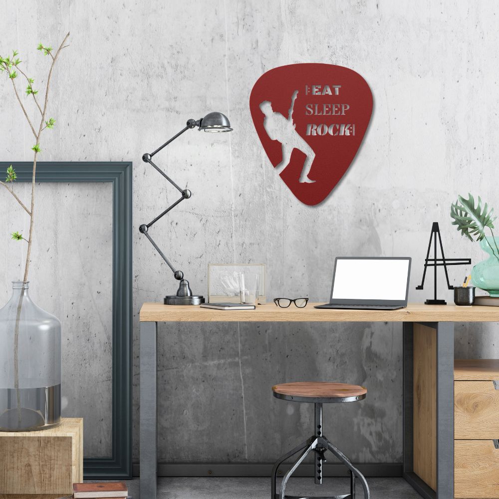 A modern workspace showcases a wooden desk with a laptop, lamp, and decorative items. On the concrete wall hangs the "Eat, Sleep, Rock, Repeat" guitar pick metal wall art made in the USA from durable powder-coated steel—the perfect gift for guitarists. A small potted plant rests on the desk.