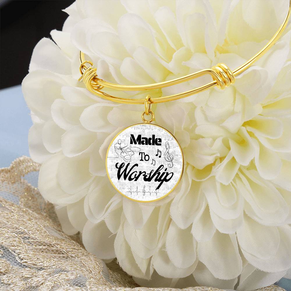 A Personalized Made to Worship Drums Round Pendant Bangle in silver, engraved with sheet music and the words “Made to Worship,” rests on a large white flower. This elegant bracelet is a thoughtful appreciation gift for worship team members or Christian drummers, with delicate lace fabric partially visible nearby.