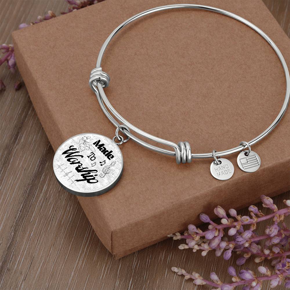 A Personalized Made to Worship Drums Round Pendant Bangle – Silver Sheet Music sits atop a brown box with sprigs of purple flowers nearby—an elegant choice for worship team appreciation or as a personalized drummer gift.