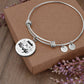 A Personalized Made to Worship Drums Round Pendant Bangle – Silver Sheet Music sits atop a brown box with sprigs of purple flowers nearby—an elegant choice for worship team appreciation or as a personalized drummer gift.