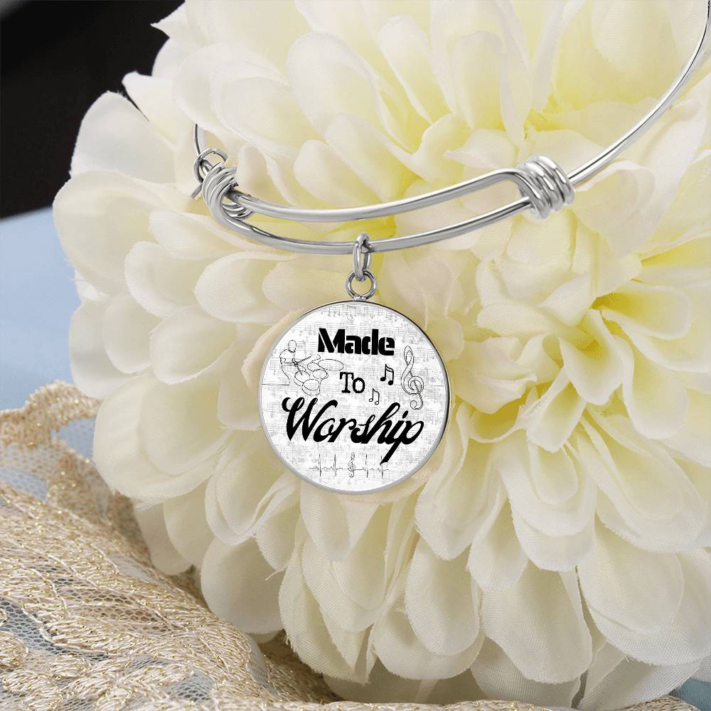 The Personalized Made to Worship Drums Round Pendant Bangle – Silver Sheet Music rests on a large, light-colored flower. This silver bracelet features a round charm inscribed with “Made to Worship,” surrounded by music notes and a treble clef, making it a thoughtful worship team appreciation or personalized drummer gift.