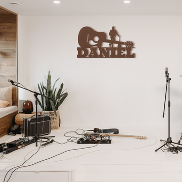 A cozy musician's corner with guitar pedals on the floor, an Acoustic Guitar leaning against a chair, and a metal wall art sign bearing the personalized name 'Daniel' with musical motifs.