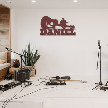 A cozy music corner featuring a personalized Acoustic Guitar with Sitting Guitarist Wall Sign Custom Name metal wall art, an acoustic guitar resting on the floor beside a pedalboard, and an amplifier in a warmly lit room with a plant adding