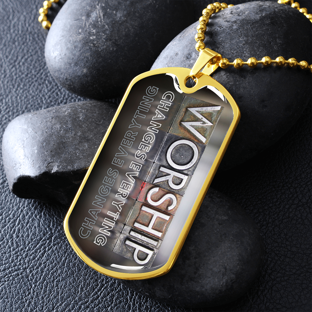 Gold military-style pendant necklace for a worshipper with bold vertical worship typography and “Worship Changes Everything”, beside black stones